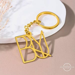 May include: A gold-colored metal keychain with the letters "DAYM" in a stylized, open design. The keychain includes a chain and a circular key ring. The background is a soft pink and white surface.