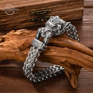 Op de afbeelding: Een zilveren armband met een wolfskop sluiting. De armband is gemaakt van een kettinglink ontwerp. De wolfskop is gedetailleerd en heeft een woeste uitdrukking.