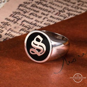 Pode incluir: Um anel de sinete prateado com uma face preta e uma letra "S" dourada estilizada. O anel está sobre um fundo castanho com escrita manuscrita e o texto "JEWEL CRAFT SIGNATURE".