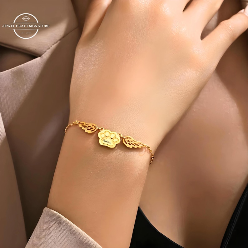 May include: Gold bracelet with a paw print charm and two angel wings charms. The paw print charm has the name "Bailey" engraved on it.