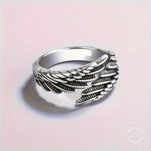 May include: A silver-colored ring with a detailed wing design. The ring features a band with a smooth, polished finish and an open design at the front, showcasing the wing's intricate feather detailing. The ring is set against a soft pink background.