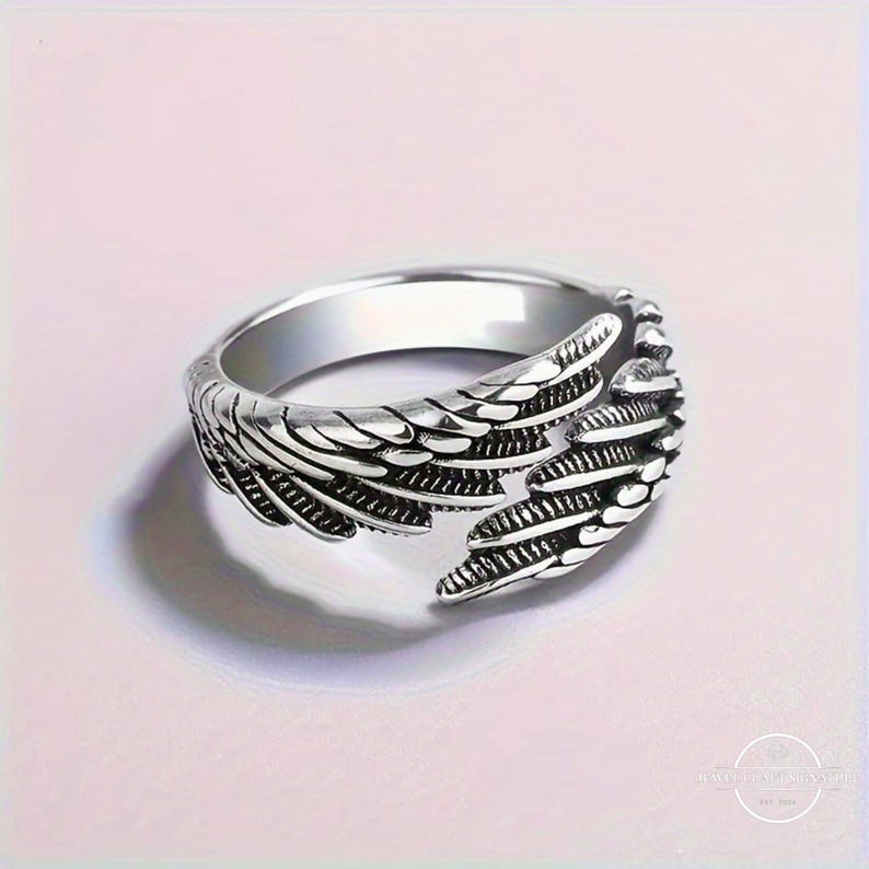 May include: A silver-colored ring with a detailed wing design. The ring features a band with a smooth, polished finish and an open design at the front, showcasing the wing's intricate feather detailing. The ring is set against a soft pink background.