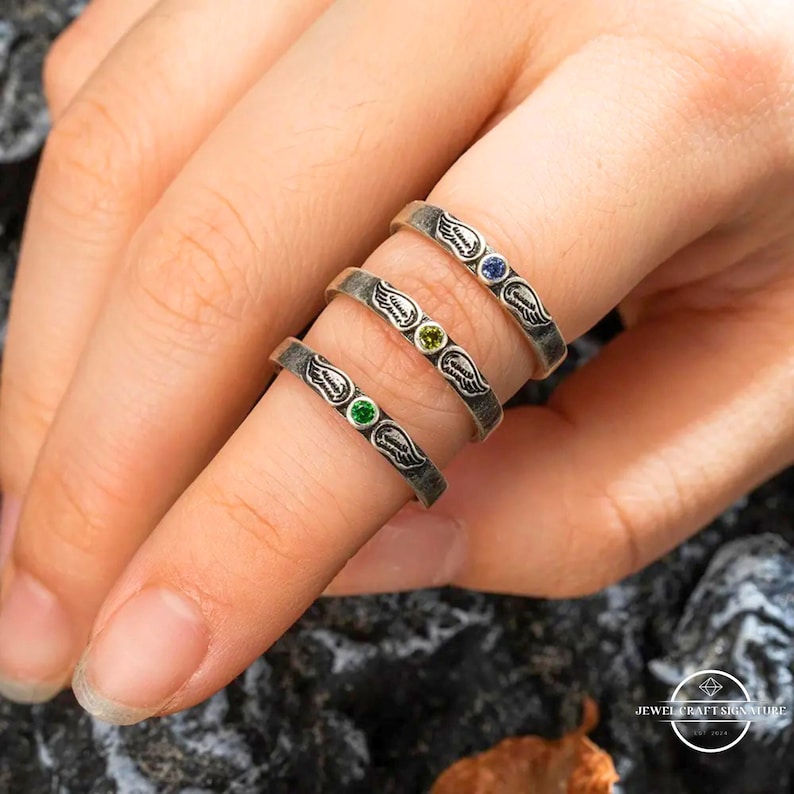 K&ouml;nnte beinhalten: Drei silberne Ringe mit Fl&uuml;gelmotiven und farbigen Edelsteinen. Die Ringe haben einen gr&uuml;nen, gelben und blauen Edelstein. Die Ringe werden an einem Finger getragen.