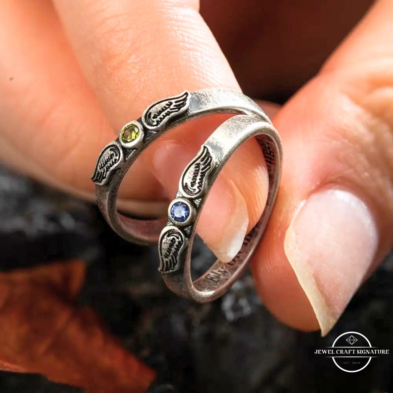 K&ouml;nnte beinhalten: Zwei silberne Ringe mit Fl&uuml;gelmotiven und Edelsteinen. Ein Ring hat einen gr&uuml;nen Edelstein, der andere einen blauen Edelstein. Die Ringe werden von den Fingern einer Person gehalten. Die Ringe haben einen Vintage- und Antik-Stil.