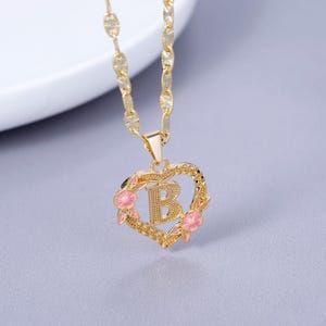 May include: Gold chain necklace with a heart-shaped pendant featuring the letter "B" surrounded by pink flowers.