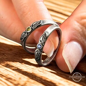 K&ouml;nnte beinhalten: Zwei silberne Ringe mit Fl&uuml;gelmotiven und Edelsteinen. Ein Ring hat einen gr&uuml;nen Edelstein, der andere einen blauen Edelstein. Die Ringe werden von den Fingern einer Person gehalten, was die komplizierten Details des Schmucks hervorhebt.