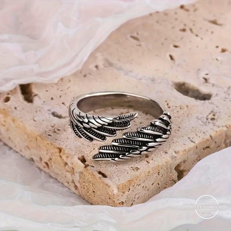 May include: A silver-colored ring with a wing design. The ring features detailed feather-like patterns, with the wings wrapping around the band. The ring is displayed on a textured, light brown surface.