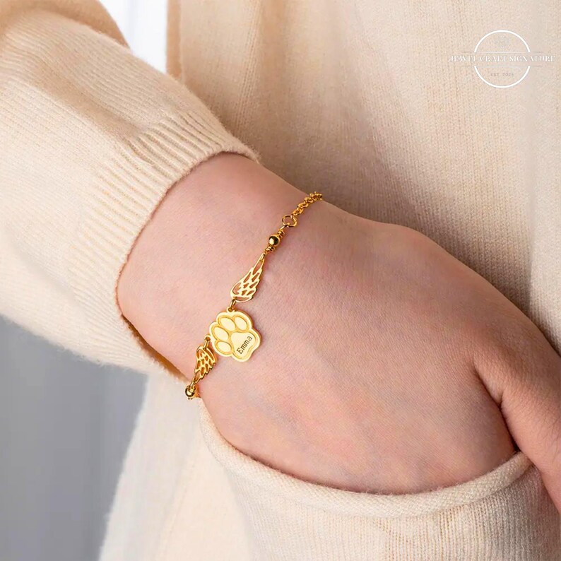May include: Gold bracelet with a paw print charm and angel wings. The charm is engraved with the name "Emma".