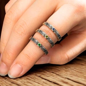 K&ouml;nnte beinhalten: Drei silberne Ringe mit einem Fl&uuml;geldesign und farbigen Edelsteinen. Jeder Ring hat einen anderen Edelstein: gr&uuml;n, gelb und blau. Die Ringe werden an einem Finger getragen und zeigen die filigranen Details des Schmucks.