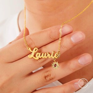 Könnte beinhalten: Eine goldfarbene Halskette mit dem Namen "Laurie" in Schreibschrift. Ein kleiner Sonnenblumenanhänger mit einem roten Stein hängt darunter. Die Halskette wird auf der Haut einer Person präsentiert.