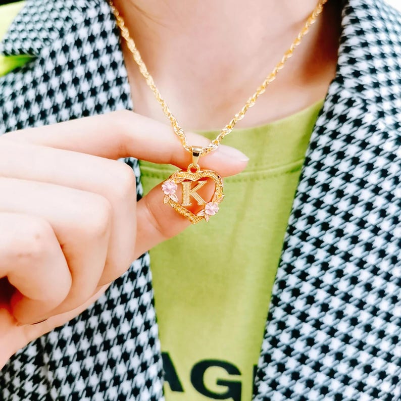 May include: Gold heart-shaped pendant necklace with a pink letter K and small pink flowers. The necklace is on a gold chain.