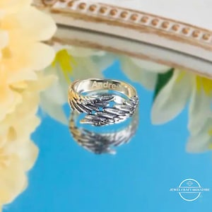 May include: A silver ring with a feather design. The ring has the name "Andre" engraved on the inside. The ring is displayed on a reflective blue surface, with a blurred floral background.