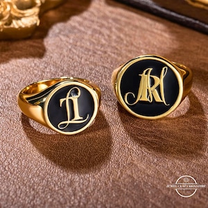 May include: Two gold signet rings with black enamel and engraved initials. The ring on the left has the initials "LI" and the ring on the right has the initials "MR".