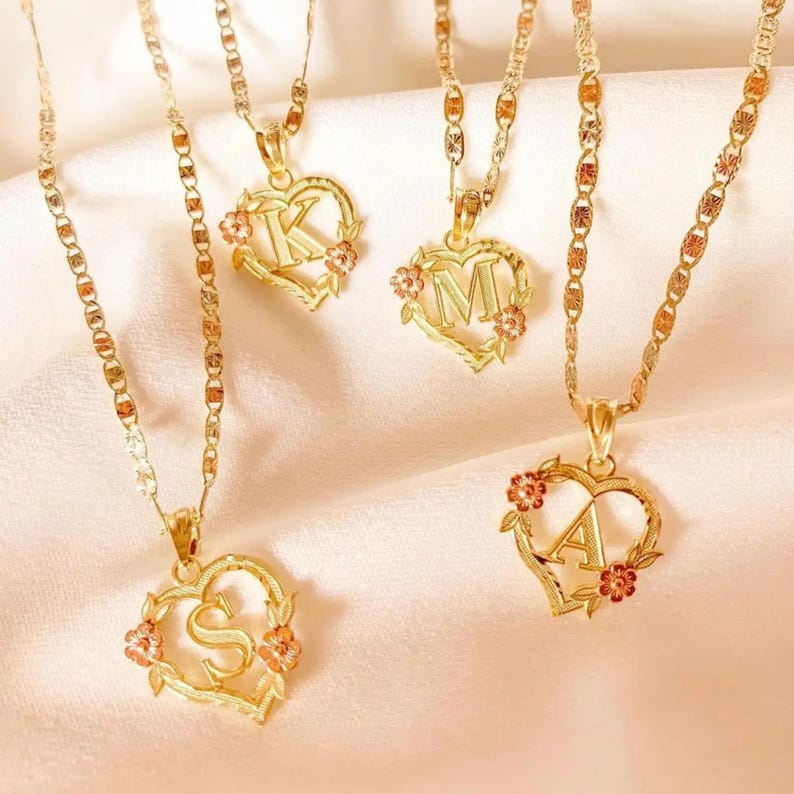 May include: Four gold necklaces with heart-shaped pendants featuring the letters K, M, A, and S. Each pendant is adorned with small pink and red flowers.
