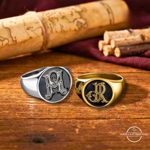 May include: Two signet rings with black faces and ornate initial designs. One ring is silver, the other is gold. The rings are on a wooden surface with a scroll and other decorative items in the background.