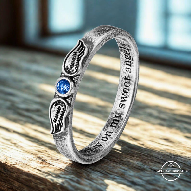 K&ouml;nnte beinhalten: Ein silberner Ring mit einem blauen Edelstein und Fl&uuml;gelmotiven. Der Ring tr&auml;gt die Inschrift "fly on my sweet angel". Der Ring liegt auf einer Holzoberfl&auml;che.