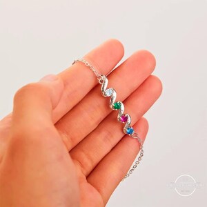 May include: A silver bracelet with a wavy design, featuring four colorful gemstones: light blue, green, magenta, and dark blue. Each stone is set in a silver setting. The bracelet is held in a hand.