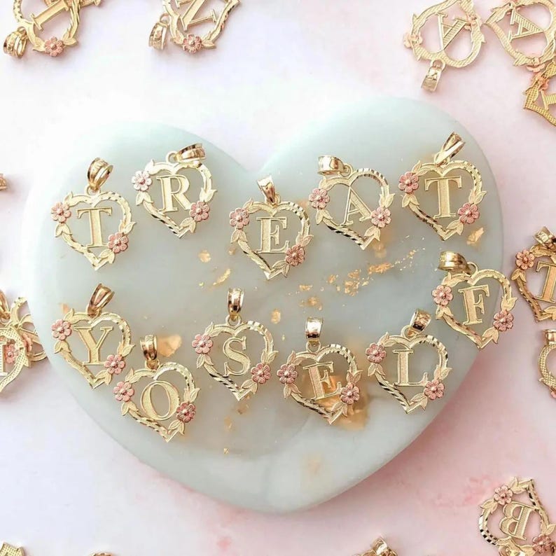 May include: A collection of gold heart-shaped pendants with different letters, each letter is surrounded by a floral design. The pendants are arranged on a white heart-shaped surface.
