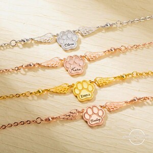 May include: Four gold, silver, and rose gold bracelets with paw print charms and angel wings. Each charm has a name engraved on it: Olivia, Keira, Emma, and Bailey.