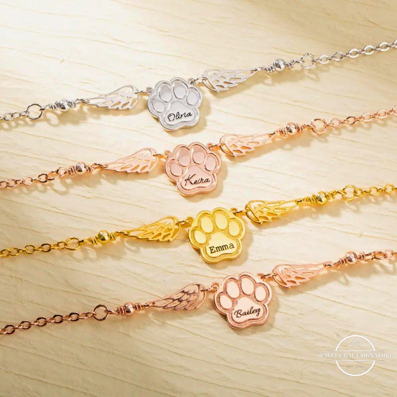 May include: Four gold, silver, and rose gold bracelets with paw print charms and angel wings. Each charm has a name engraved on it: Olivia, Keira, Emma, and Bailey.