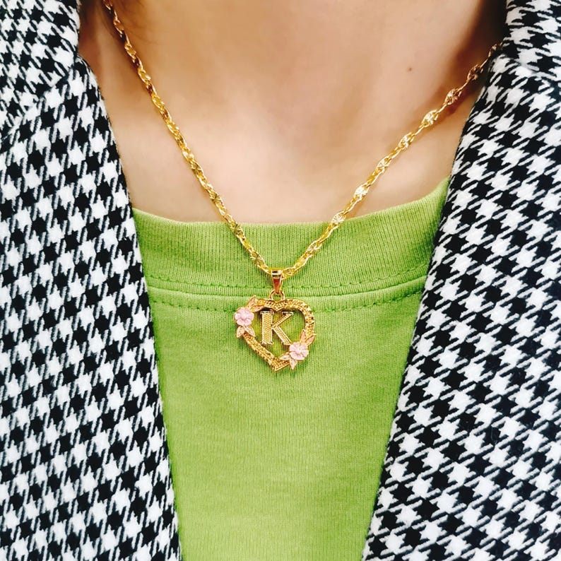 May include: A gold chain necklace with a heart-shaped pendant featuring the letter "K" surrounded by pink flowers.
