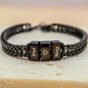 Könnte beinhalten: Schwarzes Kettenarmband mit drei schwarzen, rechteckigen Perlen. Die Perlen sind mit den Namen "Fred" und "Moxy" sowie einem Herzsymbol graviert. Das Armband liegt auf einer hellbeigen Oberfläche.