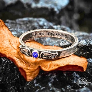 May include: A silver ring inscribed with "Fly on my sweet angel." The ring has a blue gemstone and wing motifs. It rests on a brown leaf against a dark grey stone backdrop.