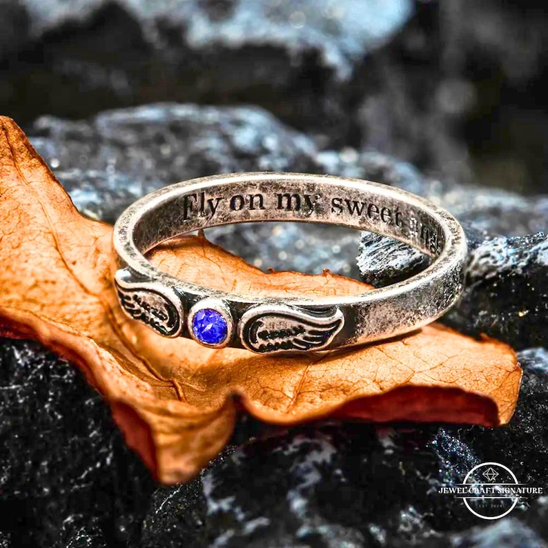 K&ouml;nnte beinhalten: Ein silberner Ring mit der Inschrift "Fly on my sweet angel". Der Ring hat einen blauen Edelstein und Fl&uuml;gelmotive. Er liegt auf einem braunen Blatt vor einem dunkelgrauen Steinhintergrund.