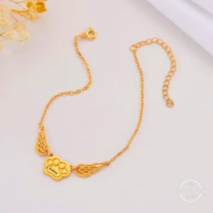 May include: Gold chain bracelet with a paw print charm and angel wings. The charm is engraved with the name "Bailey".