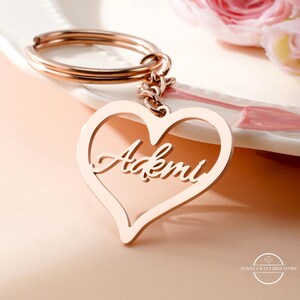 May include: Rose gold keychain with a heart-shaped pendant. The pendant features the name "Ademi" in a cursive font. The keychain includes a split ring and a chain. The background includes a white plate and pink flowers.