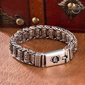 May include: Silver-toned bracelet with intricate engraved patterns and a secure clasp. The bracelet features cylindrical beads with etched symbols and a rectangular clasp with a central design. The jewelry is displayed on a brown surface.