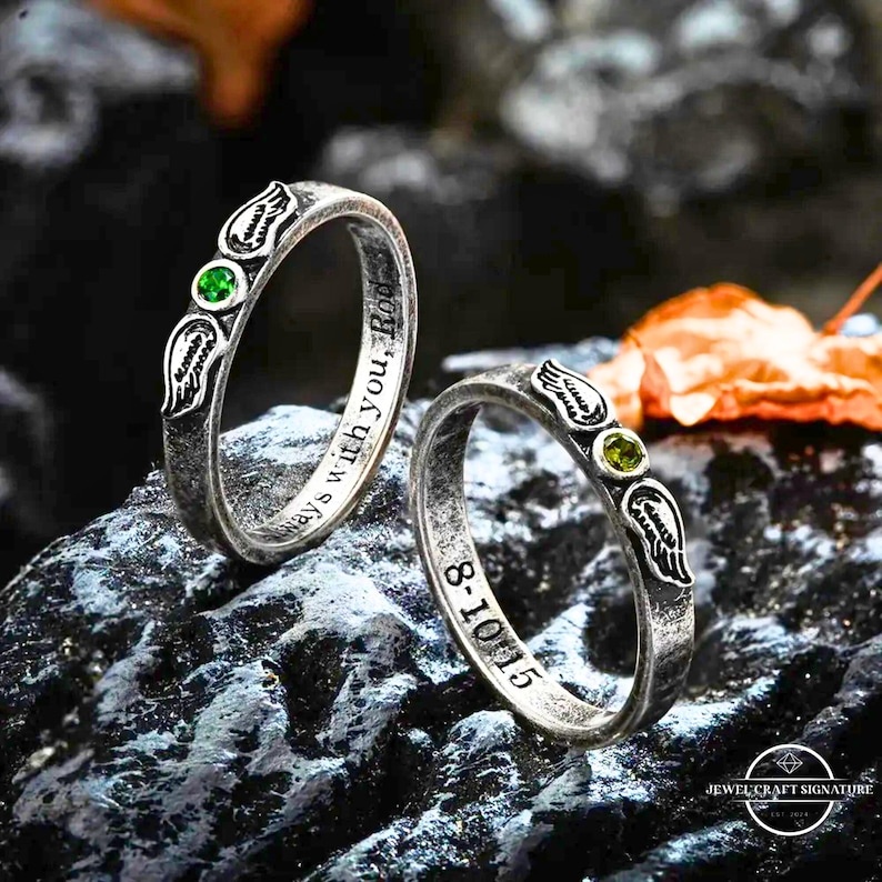 K&ouml;nnte beinhalten: Zwei silberne Ringe mit eingravierten Details und einem Edelstein. Jeder Ring hat ein Fl&uuml;geldesign und einen kleinen, runden gr&uuml;nen Edelstein. Ein Ring hat die Inschrift "always with you, Rod" und der andere "8-10-15".
