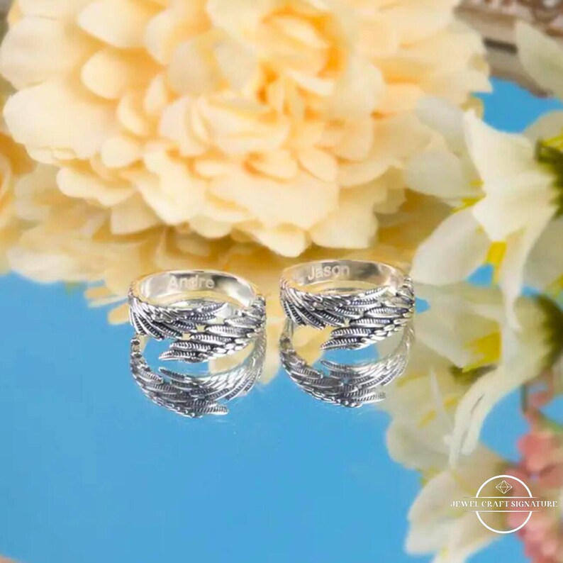 May include: Two silver rings with a feather design, each engraved with a name. One ring says "Andre" and the other says "Jason". The rings are displayed on a reflective surface with a floral background.