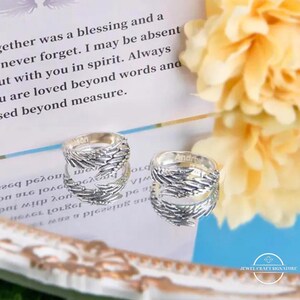 May include: Two silver rings with a feather design are displayed on a reflective surface. The rings have the names "Jason" and "Andre" engraved on them. The background includes a blurred floral arrangement and text.