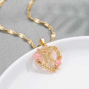 May include: Gold necklace with a heart-shaped pendant featuring the letter K surrounded by pink flowers.