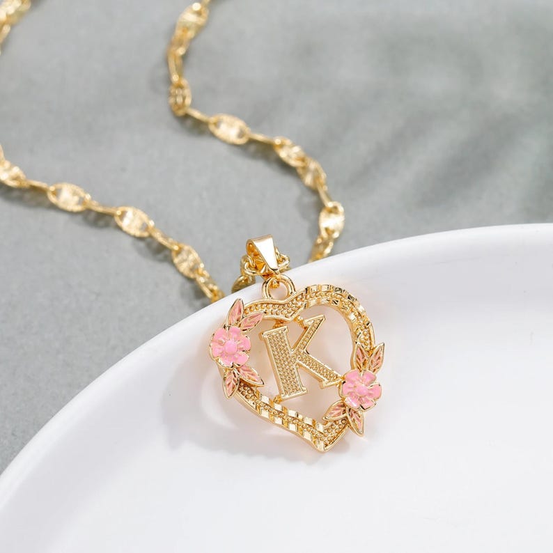 May include: Gold necklace with a heart-shaped pendant featuring the letter K surrounded by pink flowers.