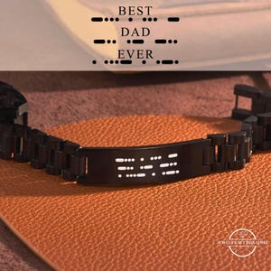 Pode incluir: Uma pulseira de metal preta com a mensagem "BEST DAD EVER" em código Morse. A pulseira está sobre uma superfície laranja texturizada. As palavras "JEWEL CRAFT SIGNATURE" são visíveis no canto inferior direito.