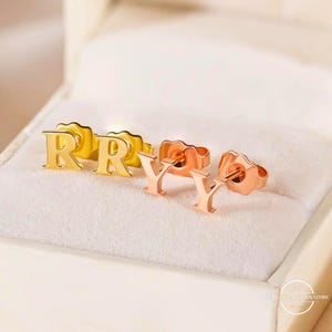 May include: Gold and rose gold letter stud earrings. The earrings are in the shape of the letters "R" and "Y". The earrings are displayed in a white jewelry box.