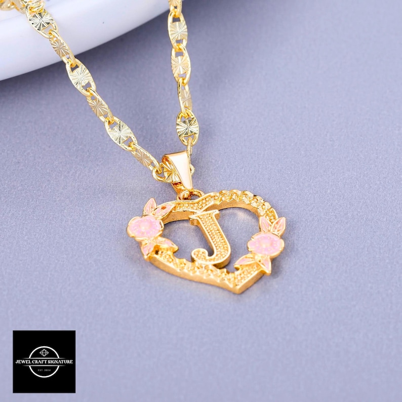 May include: Gold chain necklace with a heart-shaped pendant featuring the letter J and pink flower accents.