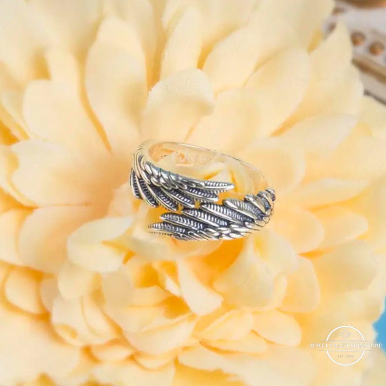 May include: A silver-toned ring with a feather design. The ring is set against a soft yellow background. The ring's design features detailed feather patterns, creating a textured appearance. The ring is a piece of jewelry.