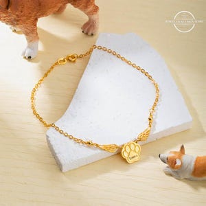 May include: Gold chain bracelet with a paw print charm and angel wings. The charm is engraved with the name "Emma".
