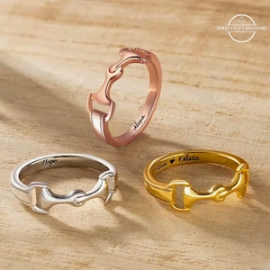 May include: Three rings in rose gold, silver, and gold, each featuring a horse bit design. The rose gold ring is engraved with "Alexa", the silver ring with "Hope", and the gold ring with "Kristan ♥ Olivia". The rings are displayed on a light brown wooden surface.