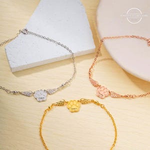 May include: Three silver, rose gold, and gold bracelets with paw print charms and angel wings. Each charm is engraved with a name: "Olivia", "Bailey", and "Emma".