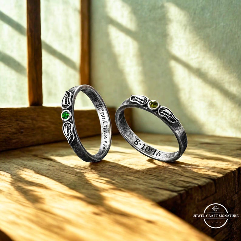 K&ouml;nnte beinhalten: Zwei silberne Ringe mit einem Used-Look-Finish. Jeder Ring hat ein Edelstein- und Fl&uuml;geldesign. Ein Ring hat einen gr&uuml;nen Edelstein, der andere einen gelb-gr&uuml;nen Edelstein. Ein Ring ist mit dem Text "always with you, Babe" graviert.