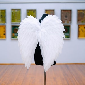 Puede incluir: Alas de ángel blancas en un maniquí negro de pie contra una pared con obras de arte enmarcadas.
