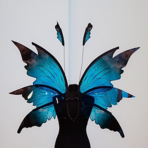 Könnte beinhalten: Eine schwarze Schaufensterpuppe mit großen blauen Schmetterlingsflügeln, die am Rücken befestigt sind. Die Flügel haben einen schwarzen Umriss und eine glitzernde blaue Oberfläche.