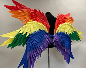 May include: Rainbow-colored costume wings displayed on a black mannequin. The wings are arranged in a layered design, with vibrant hues of red, orange, yellow, green, blue, and purple. The wings are made of a material that appears to be fabric or paper.