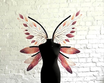 May include: A black dress form with decorative fairy wings. The wings feature a black frame with branches and leaves in shades of red, orange, and pink. The dress form is on a metal stand against a white brick wall.
