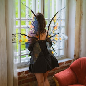 May include: A person wearing a black dress and fairy wings with yellow and orange floral accents. The wings are transparent with black frames. The person is standing near a window with a red armchair in the background.