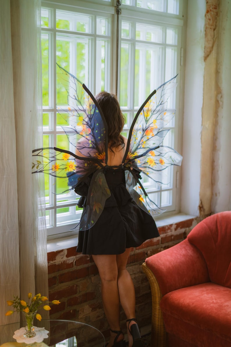May include: A person wearing a black dress and fairy wings with yellow and orange floral accents. The wings are transparent with black frames. The person is standing near a window with a red armchair in the background.
